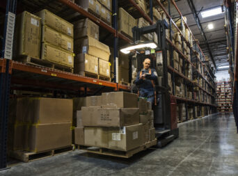 Warehouse worker wearing a safety harness while opeating a motorized stock picker in an aisle between large racks of cardboard boxes holding product on pallets in a large distribution warehouse.